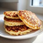 A close-up stack of four golden-brown, fluffy almond flour pancakes served on a white plate near a window.