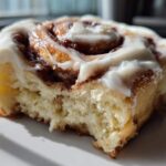 A close-up, cross-section view of a fluffy, homemade cinnamon roll topped with thick white frosting.
