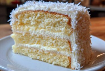 Close-up of a fluffy slice of coconut cake layered with white frosting and covered in shredded coconut.