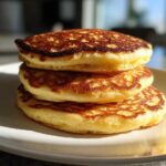 A stack of three golden-brown coconut flour pancakes sitting on a white plate, lit by sunlight.