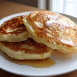 A stack of three fluffy discard pancakes drizzled generously with golden syrup on a white plate.