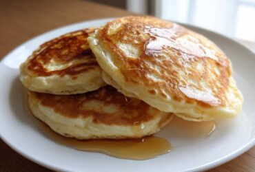 A stack of three fluffy discard pancakes drizzled generously with golden syrup on a white plate.