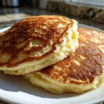 A close-up, sunlit stack of three fluffy greek yogurt pancakes with golden-brown, lacy tops on a white plate.