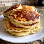 A close-up of a stack of four fluffy pancakes drizzled with syrup, showcasing their golden brown tops.
