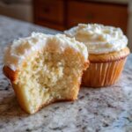 Close-up of a vanilla cupcake cut in half showing its fluffy texture, topped with white frosting, next to a whole vanilla cupcake.