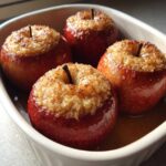 Four glossy, red baked apple desserts topped with a golden, crunchy brown sugar filling, sitting in a white baking dish.