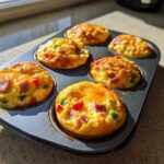 Six golden-brown Egg Muffin Cups with ham and peppers baked in a dark muffin tin.