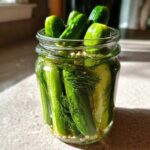 A glass jar filled with fresh cucumbers, dill, and spices for making refrigerator pickles, sitting in sunlight.