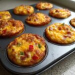 Close-up of several golden-brown Egg Muffin Cups baked in a dark muffin tin, topped with melted cheese and visible peppers.