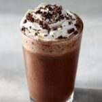 A tall glass filled with rich, blended frozen hot chocolate topped with whipped cream and chocolate shavings.