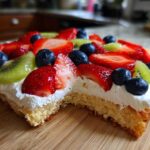 Close-up of a slice of fruit pizza sugar cookie topped with whipped cream, strawberries, blueberries, and kiwi.
