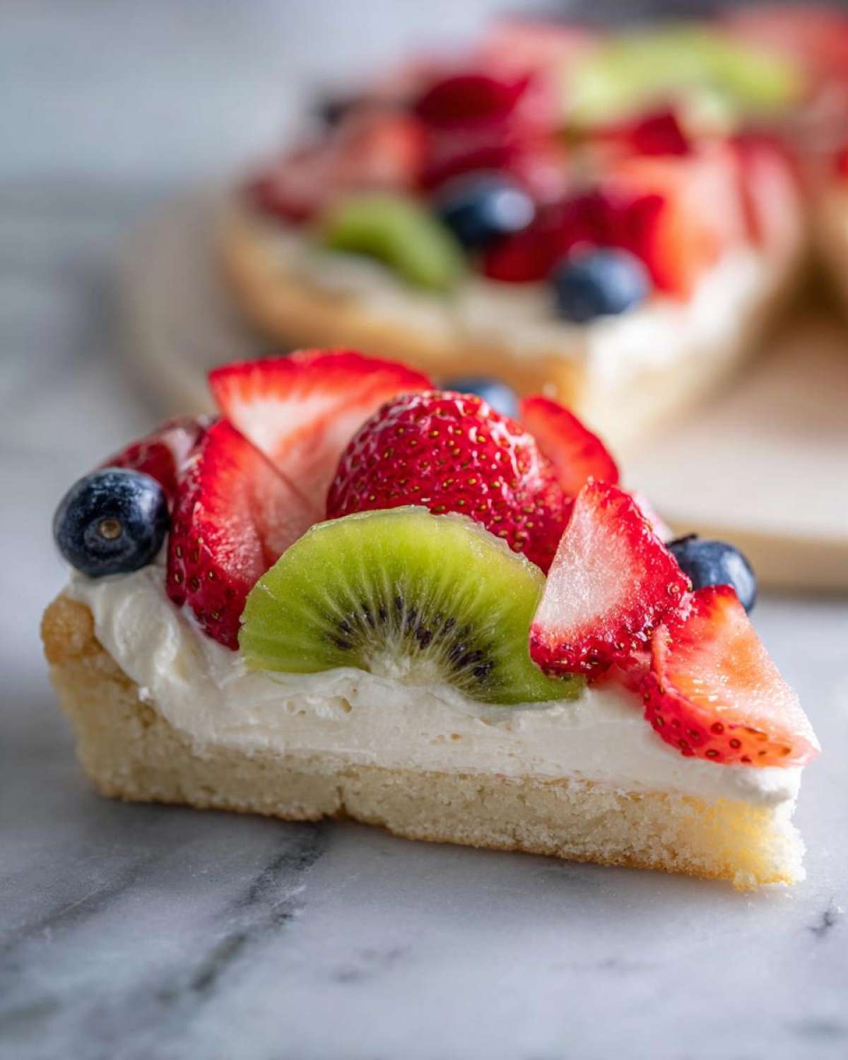 Close-up of a slice of fruit pizza sugar cookie topped with cream cheese frosting, strawberries, blueberries, and kiwi.