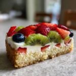 Close-up of a slice of fruit pizza sugar cookie topped with cream cheese frosting, strawberries, kiwi, and blueberries.