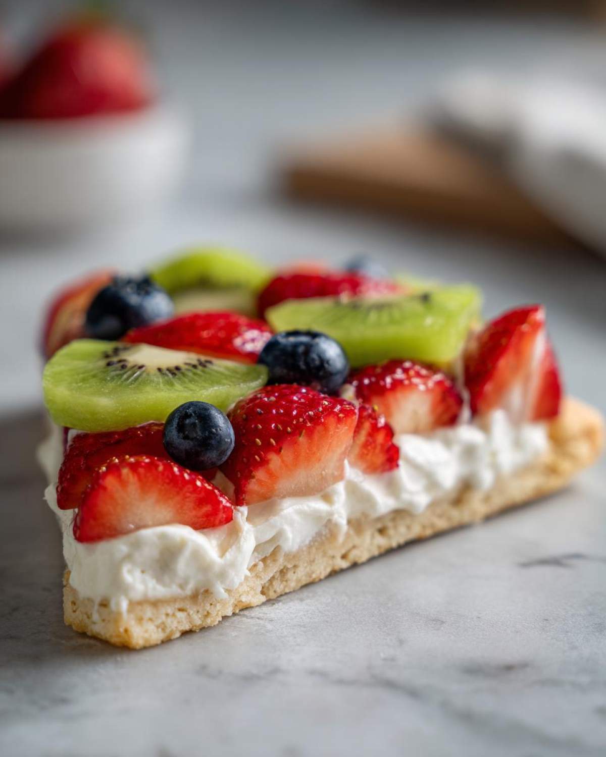 A close-up slice of fruit pizza sugar cookie topped with white frosting, strawberries, blueberries, and kiwi slices.