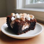 A fudgy piece of hot chocolate brownies topped with toasted mini marshmallows on a white plate.