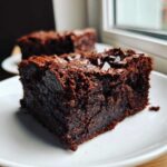 Close-up of a fudgy, dark chocolate slice of sweet potato brownies topped with chocolate chips on a white plate.