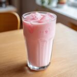 A tall glass filled with pink strawberry milk and ice cubes sitting on a light wooden table.