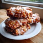 A stack of four golden-brown, freshly fried apple fritter pastries covered in a sweet white glaze.