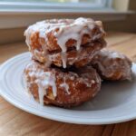 Close-up of three golden brown, irregularly shaped apple fritter donuts stacked on a white plate, drizzled with white glaze.