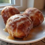 Three golden brown baked apple fritters drizzled with a thick white glaze, resting on a white plate.