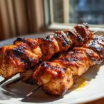 Close-up of two perfectly grilled, glazed chicken skewers resting on a white plate, featuring charred edges.