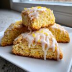 Close-up of four triangular lemon scones stacked, drizzled with white glaze and topped with bright yellow lemon zest.