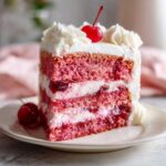 A tall, moist slice of pink cherry cake layered with white frosting and cherry filling, topped with whipped cream and a maraschino cherry.