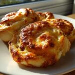 Close-up of freshly baked sausage pinwheels with melted, golden-brown cheese topping, served on a white plate.