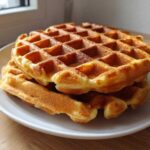 A stack of golden brown, freshly made high protein waffles resting on a white plate.
