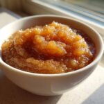 Close-up of rich, brown homemade applesauce sprinkled with spices, served in a white bowl near a sunny window.