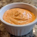 Close-up of creamy, orange-hued big mac sauce speckled with spices, served in a small white dipping bowl.
