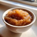 Close-up of homemade applesauce, rich brown in color and sprinkled with cinnamon, served in a small white bowl.
