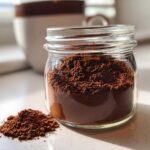 A glass jar filled with rich brown homemade hot cocoa mix, with a small pile spilled next to it.