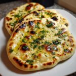 Close-up of three pieces of freshly cooked homemade naan bread, brushed with melted butter and topped with chopped green herbs.