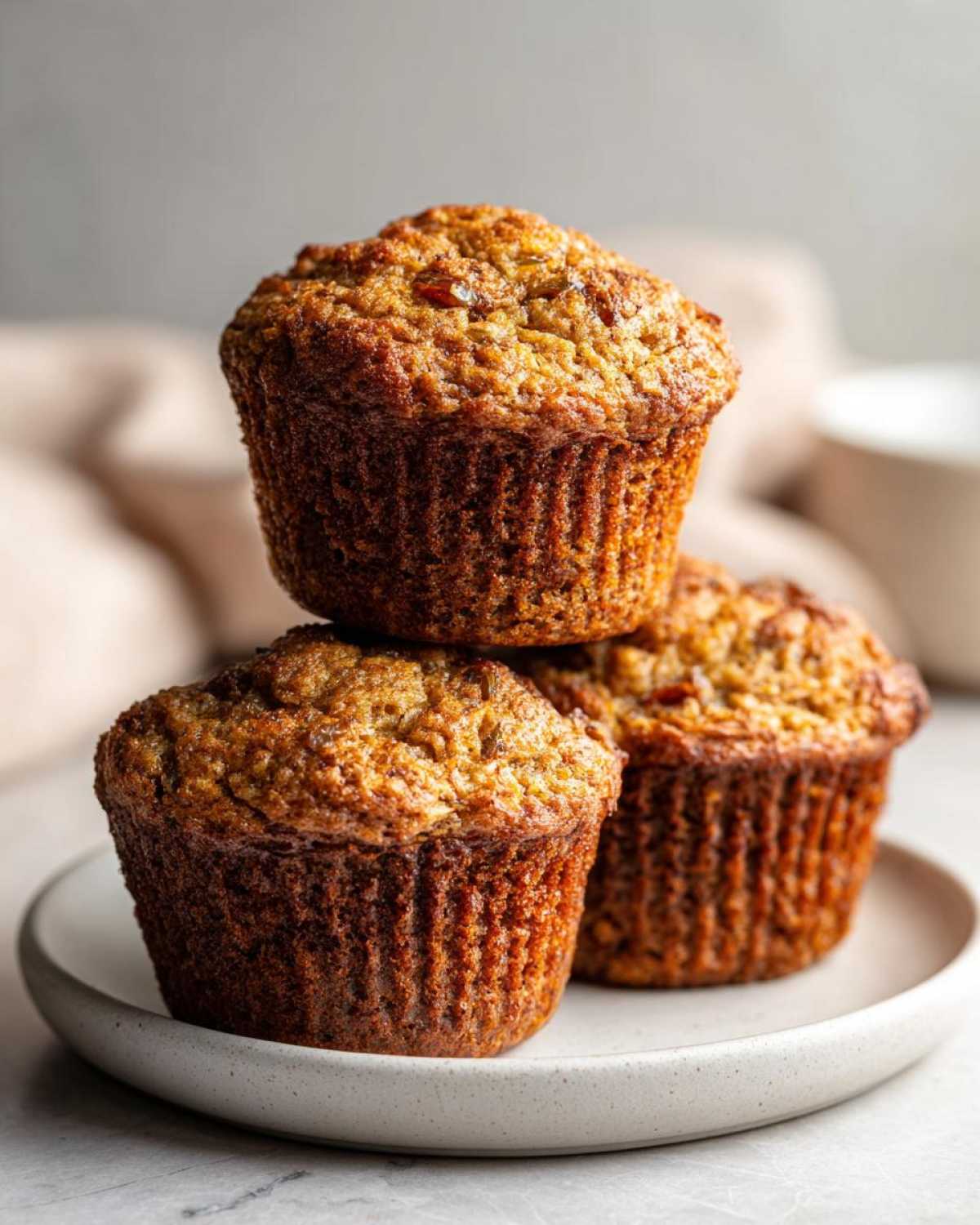 Three golden brown honey bran muffins stacked on a small, light-colored plate.