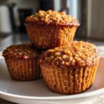 Three golden brown honey bran muffins stacked on a white plate, featuring a textured, slightly sticky top.