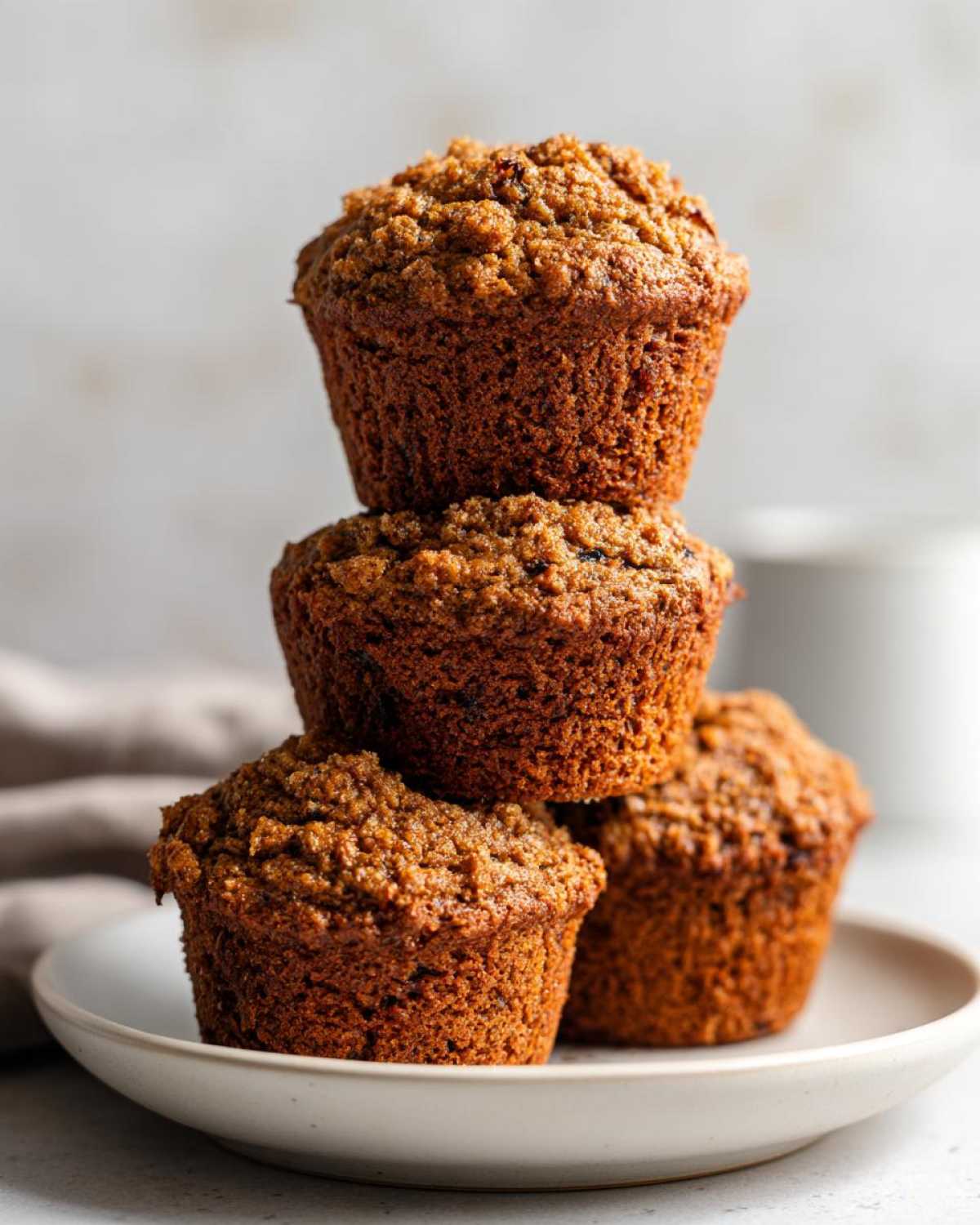 A stack of four golden-brown, textured honey bran muffins sitting on a light ceramic plate.