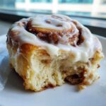 A close-up of a fluffy, iced cinnamon roll with a bite missing, showing the soft interior and cinnamon swirl.