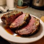 Three thick, perfectly cooked slices of prime rib roast with a dark herb crust resting in au jus on a white plate.
