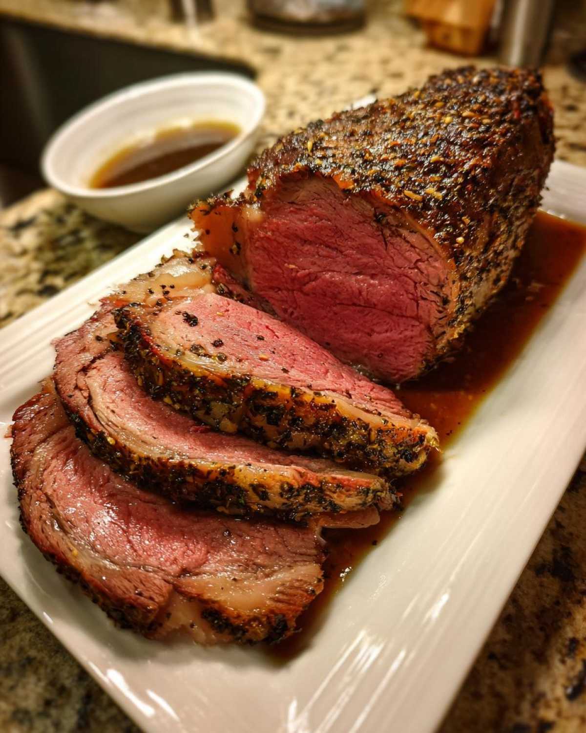 Close-up of a juicy, medium-rare prime rib roast, sliced and resting in its au jus on a white platter.