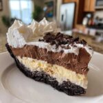 A tall slice of layered ice cream pie with a dark cookie crust, vanilla, chocolate mousse, and whipped cream topping.