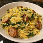 Close-up of a white bowl filled with creamy lemon garlic parmesan orzo pasta mixed with sausage slices and spinach.