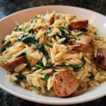 Close-up of a white bowl filled with creamy Lemon Garlic Parmesan orzo pasta mixed with spinach and sliced sausage.