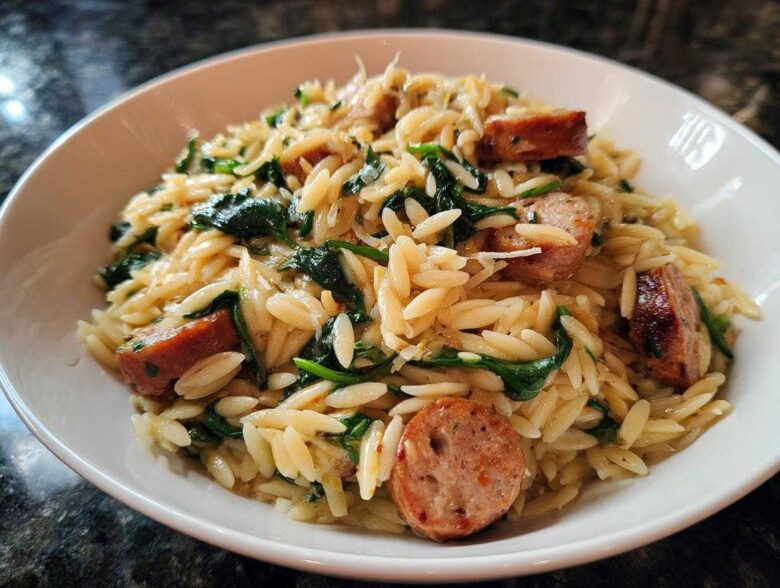 Close-up of a white bowl filled with creamy Lemon Garlic Parmesan orzo pasta mixed with spinach and sliced sausage.