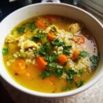 Close-up of a white bowl filled with Lemon Ginger Turmeric Chicken and Rice Soup, featuring bright yellow broth, rice, carrots, and parsley.
