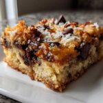 A close-up view of one square of rich magic cookie bars on a white plate, showing melted chocolate and coconut topping.