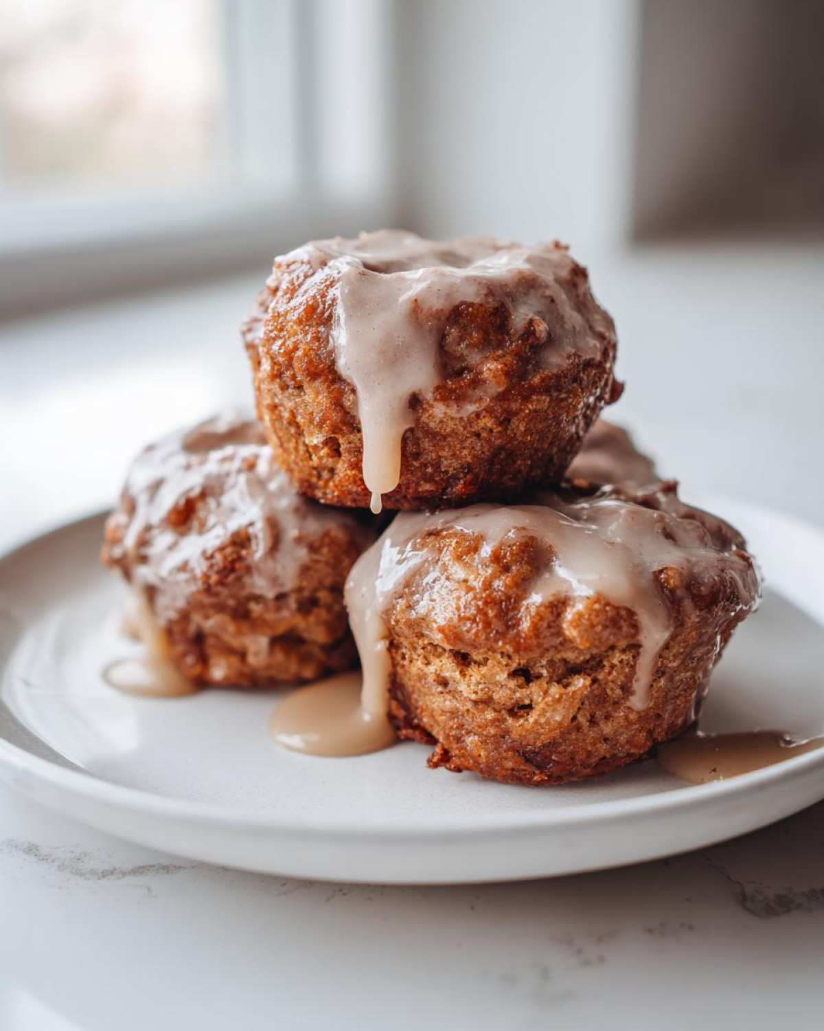 Three golden brown baked apple fritters stacked on a white plate, generously drizzled with a light brown glaze.