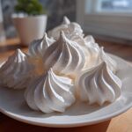 A close-up of several bright white, perfectly piped meringue cookies piled on a white plate.