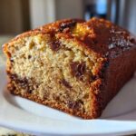 Close-up of a moist slice of chocolate chip banana bread with a dark, caramelized crust on a white plate.