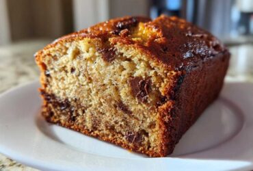 Close-up of a moist slice of chocolate chip banana bread with a dark, caramelized crust on a white plate.